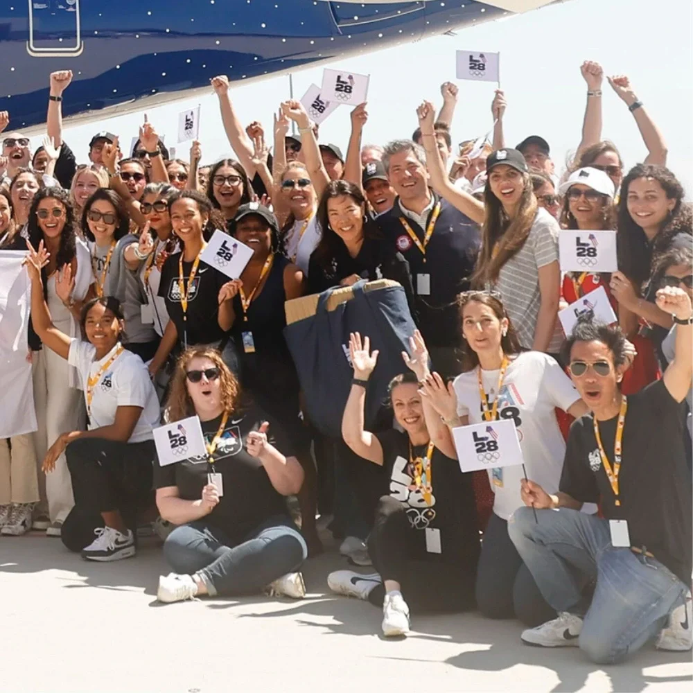 A group of LA28 employees holding small LA28 flags with the Olympic rings on them in front of the Delta Olympic Flag Flight plane