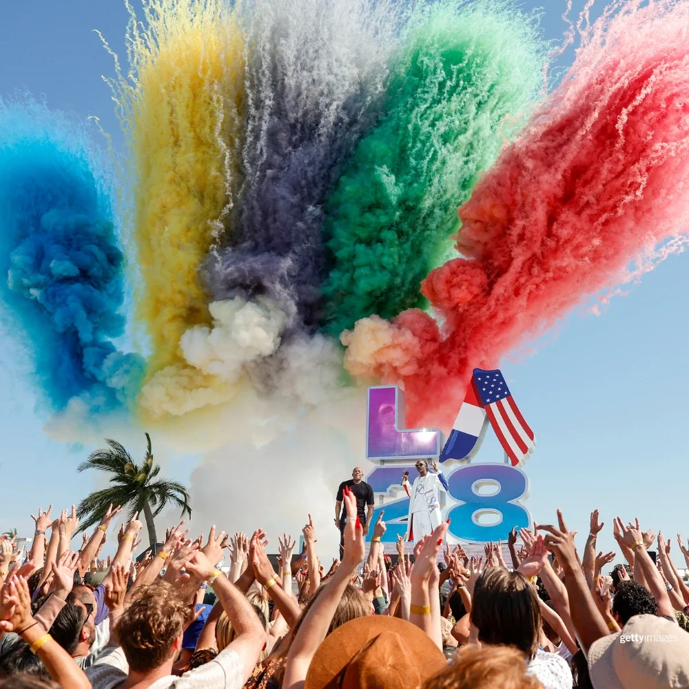 Photo of colored smoke in black, blue, red, yellow and green in front of a stage with Dr. Dre and Snoop Dogg singing at the LA28 Olympic Handover reveal ceremony
