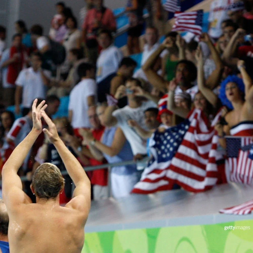 photo of a swimmer with his back to the camera clapping to fans in the stand who are waving American flags